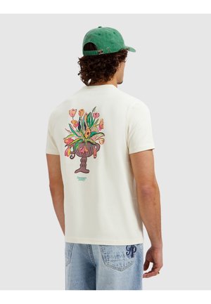 Man wearing a white t-shirt with a colorful floral vase print on the back, paired with light blue jeans and a green baseball cap.