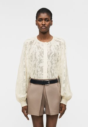 Vrouw draagt een doorschijnende crèmekleurige kanten blouse met knopen en lange mouwen, en beige shorts met hoge taille en een zwarte riem, staand tegen een witte achtergrond.