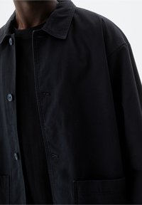 Black button-up jacket with a collared neckline, oversized fit, front pockets, and smooth texture. Close-up on fabric and button details.