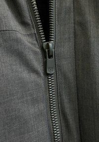 Melna auduma virsma ir metāla rāvējslēdzis ar vilkšanas cilpiņu, blakus ir plakana šuve, kas demonstrē izturīgu, teksturētu virsmu.