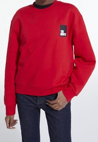 Sudadera roja con cuello redondo, puños y cintura acanalados, con un logotipo rectangular negro en el área del pecho izquierdo.