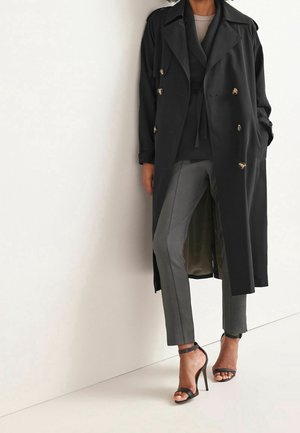 Personne portant un long manteau noir, un pantalon gris, des talons hauts noirs avec bride à la cheville, debout contre un mur blanc.