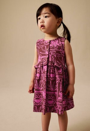 Young girl with dark hair in a side ponytail wearing a sleeveless purple and pink floral patterned dress, standing against a beige background.