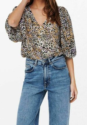 Femme portant un chemisier à manches bouffantes imprimé léopard, rentré dans un jean bleu taille haute, debout devant un fond blanc.