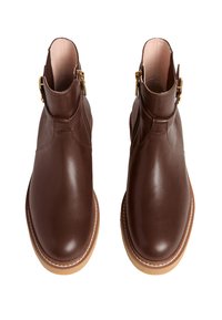 Para brązowych skórzanych botków za kostkę z jasnobrązowymi podeszwami, złotymi zamkami błyskawicznymi po bokach i klamrami, przedstawionych z widoku z góry na białym tle.