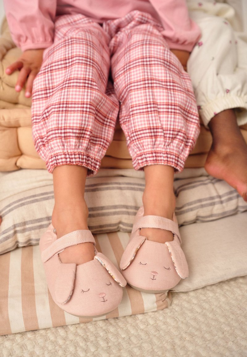 Enfant portant un pantalon à carreaux roses et des chaussons lapin roses assis sur des coussins avec un autre enfant à proximité.