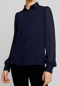 Blouse bleu marine à boutons, avec de longues manches bouffantes transparentes, un col classique et une lisière droite. Tissu doux et léger avec des détails minimalistes.