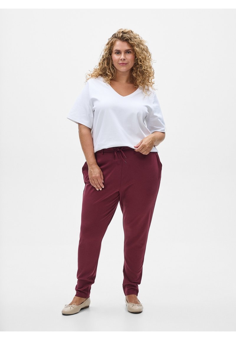 Krøllet kvinde står iført en hvid kortærmet V-hals top, bordeaux snøre bukser og beige flade sko mod en ensfarvet baggrund.