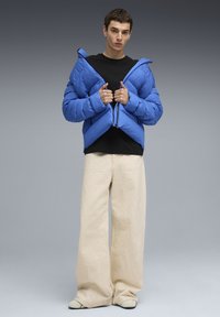 Chaqueta de abrigo azul con cuello alto y cremallera de doble sentido, llevada sobre una camisa negra. Pantalones de pierna ancha color crema completan el atuendo.