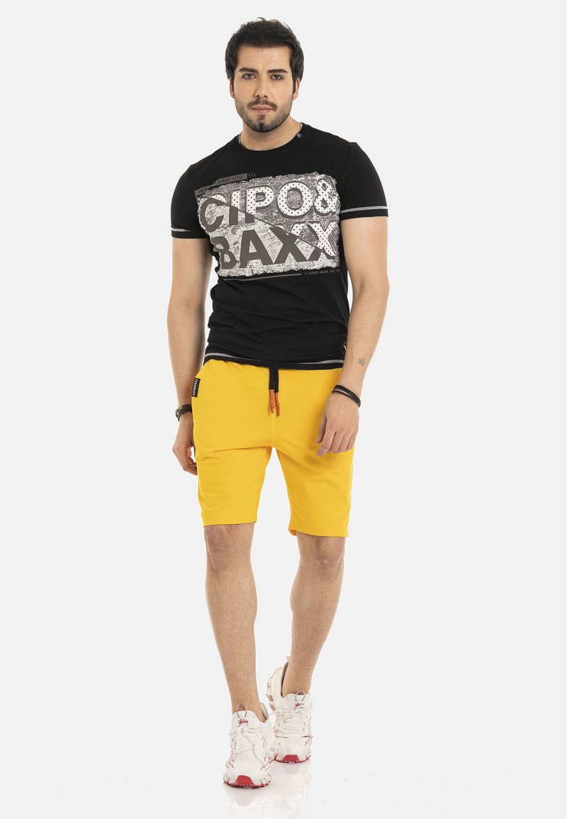 Zwart T-shirt met korte mouwen met een grafische print, gecombineerd met gele shorts en witte sneakers met rode accenten.