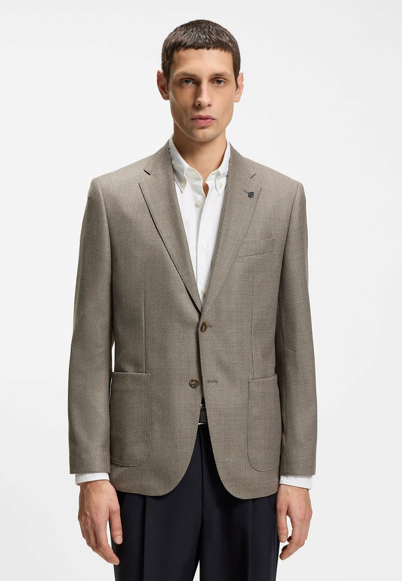 Homme portant un blazer beige texturé sur une chemise blanche boutonnée et un pantalon noir, debout devant un fond blanc uni.