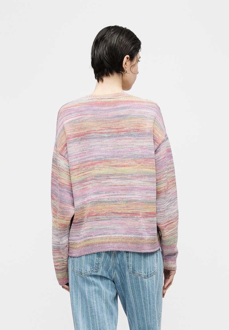 Persona con capelli corti e scuri che indossa un maglione a righe multicolori e jeans a righe blu, in piedi di spalle contro uno sfondo semplice.