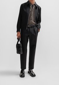 Zwarte jas over een patroonrijk overhemd, op maat gemaakte zwarte broek, glanzende zwarte schoenen en een kleine zwarte tas. Belangrijkste kleuren: zwart, bruin en beige.