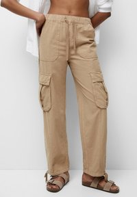 Pantalons cargo beiges avec une taille élastique, cordon de serrage, et deux grandes poches latérales ; associés à des sandales marron. Tissu en coton lisse.
