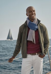 Hombre vestido con una chaqueta de ante verde, un suéter burdeos y pantalones blancos. Lleva una bufanda de punto a rayas en azul y gris. Barco de vela en el fondo.