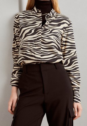 Femme portant une chemise boutonnée zébrée noire et blanche sur un col roulé noir, associée à un pantalon noir taille haute avec des poches latérales.