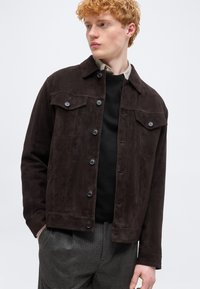 Νέος άνδρας με σγουρά κόκκινα μαλλιά, φορώντας ένα σκούρο καφέ σακάκι με κουμπιά πάνω από ένα μαύρο πουλόβερ και γκρι καρό παντελόνι, κοιτάζοντας πλάγια.