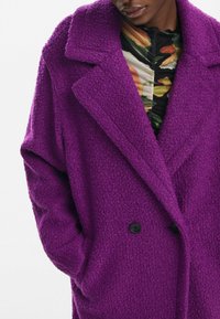 Abrigo púrpura de gran tamaño hecho de tejido texturizado. Presenta un cuello con muesca, botones negros de doble botonadura y bolsillos laterales.