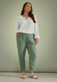 Witte textuurblouse met een strikdetail bij de hals, gecombineerd met lichtgroene, loszittende broek en witte sandaaltjes met bandjes.
