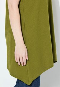 Top verde oliva con una vestibilità comoda, maniche corte, orlo arrotondato, texture morbida e cuciture visibili lungo i bordi, abbinato a jeans blu.