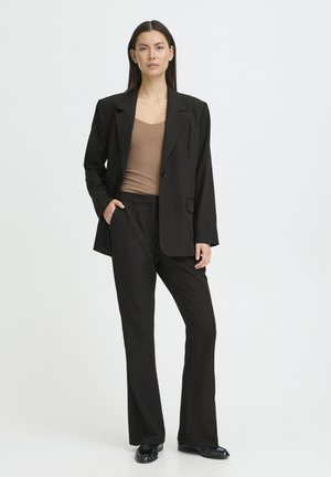 Schwarzer, maßgeschneiderter Blazer mit strukturierten Schultern, getragen über einem taillierten, hellbraunen Oberteil, kombiniert mit passenden schwarzen, geraden Hosen und Loafers.