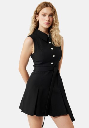 Vrouw met golvend blond haar die een mouwloze zwarte jurk draagt met kraag, decoratieve witte knopen en een tailleband, staand tegen een effen achtergrond.