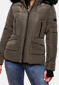 Vrouw in een olijfgroene gewatteerde jack met een rits aan de voorkant, zwarte ritszakken op de borst en een bontomrand capuchon, gecombineerd met blauwe jeans.