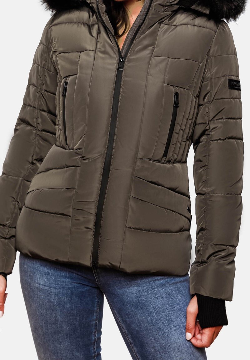 Vrouw in een olijfgroene gewatteerde jack met een rits aan de voorkant, zwarte ritszakken op de borst en een bontomrand capuchon, gecombineerd met blauwe jeans.