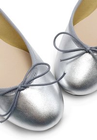 Ballerine argento con texture metallica, punta rotonda e un piccolo fiocchetto grigio davanti. L'interno presenta una fodera beige.