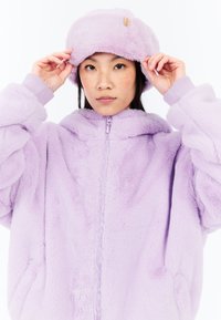 Lila tekoturkki takki, jossa on vetoketjukiinnitys, ylisuuri malli ja ribatut hihansuut. Samasta lila tekoturkista valmistettu hattu, joka on päässä, ja jossa on etiketti.