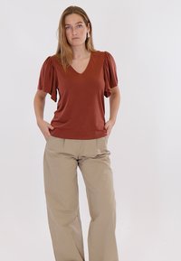 Naine seisab käed taskutes, seljas roostevärvi punnilõhisevarrukatega pluus ja beežid laia säärega püksid, taust on lihtne.