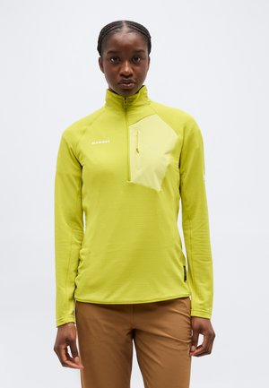 Žena v limetkovo zelenom fleece polozip svetri s náprsní vreckom, stojaca pred jednoduchým svetlým pozadím, čelom vpred.