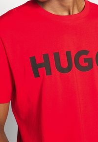 Camiseta de algodón roja con un gran logo negro "HUGO" en la parte frontal. Mangas cortas y un diseño clásico de cuello redondo.