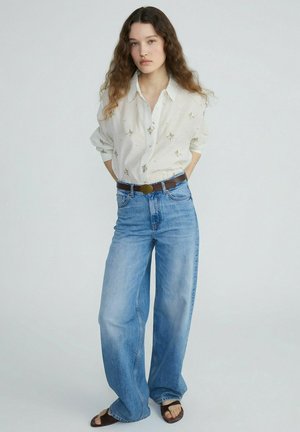 Femme aux longs cheveux bouclés portant un chemisier blanc brodé, un jean bleu large, une ceinture marron et des sandales ouvertes, debout les mains dans le dos.