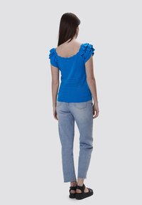 Blaues Stricktop mit Rüschensaum, gewelltem Ausschnitt, strukturierten Mustern, kombiniert mit hellblauen Jeans und schwarzen Sandalen.