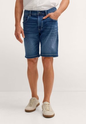 Mann trägt mittelblaue Jeansshorts, weiße Sneakers und ein weißes Hemd, mit einer Hand in der Tasche, stehend vor einem schlichten hellen Hintergrund.