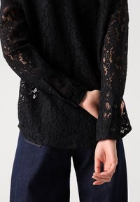 Black lace blouse with long sleeves, detailed floral pattern, buttoned cuffs, paired with dark denim trousers. Delicate, textured fabric.