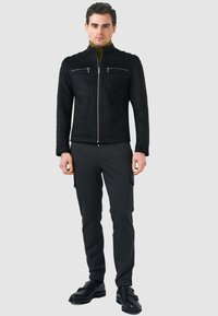 Giacca in suede nera con colletto alto, dotata di due tasche con zip sul petto e pannelli laterali a coste. Abbinata a pantaloni cargo grigio scuro e scarpe nere.