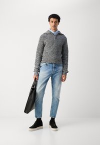 Pull en maille gris avec un col, chemise à boutons bleu clair en dessous, jeans bleu délavé, baskets noires et sac fourre-tout noir.