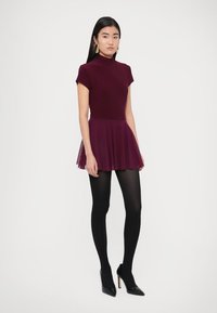 Obleka bordo barve z oprijetim zgornjim delom in razširjeno tilasto krilo. Kombinirana s črnimi hlačnimi nogavicami in visokimi petami. Gladek material in elegantna silhueta.