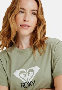Olivově zelené bavlněné tričko s velkým bílým srdcem s geometrickým vzorem a nápisem "ROXY" černým písmem dole.