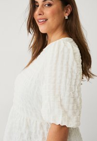 Weiße Strukturbluse mit Puffärmeln, die ein gerafftes Design aufweist. Der weiche, zerknitterte Stoff sorgt für einen leichten Look.