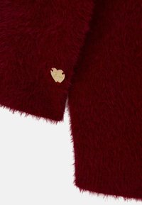 Burgundy puha pulóver, aranyszínű szív alakú fém címkével, amelyen a márkaneve látható, és egy kis strasszkő díszíti.