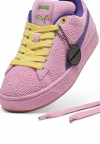 Pink sneakers med tekstureret overdel, gule snørebånd og lilla accenter. Inkluderer en sort charm og en pink snor, der hviler ved siden af.