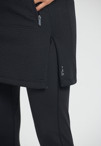 Musta aktiivivaatetus, jossa on tekstuuripinta ylhäällä, vetoketju, sivuhalkiot ja tiukat leggingsit. Sileä materiaali, virtaviivainen muotoilu ja minimalistinen brändäys.