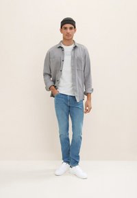 Afslappet outfit med en lys grå skjorte med knapper, hvid t-shirt, blå jeans og hvide sneakers, samt en mørk strikhue.