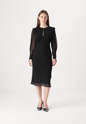 Woman wearing a black midi dress with sheer long sleeves and keyhole neckline, paired with black pointed-toe heels, standing on a white floor.