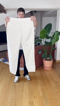 Bílé bavlněné kalhoty s elastickým pasem, rovný střih, drží je osoba v jasném vnitřním prostoru, v pozadí rostliny.