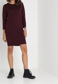 Vestido de punto burdeos con cuello redondo y mangas tres cuartos, de corte holgado, combinado con botas de tobillo negras con cordones.