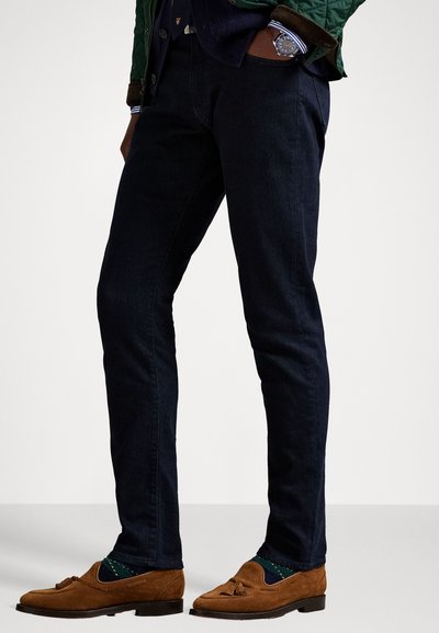 Hombre vistiendo jeans oscuros ajustados, mocasines de ante marrones con borlas, calcetines verdes de rombos y una chaqueta verde con las manos en los bolsillos.